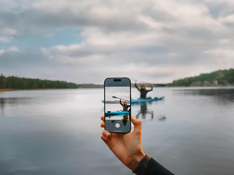 En hånd som bruker kamerakontrollen på en iPhone 16 Pro, med en person i en kano i bakgrunnen