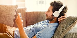 Mann ligger på sofa med smarttelefon i hånden og headset på hodet